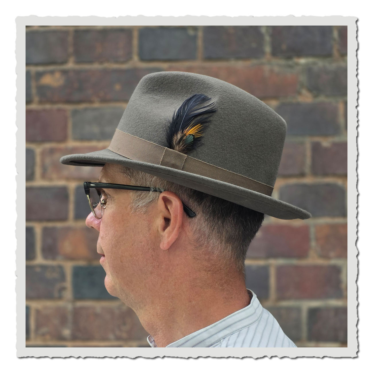 Seitenansicht des Fedora-Hutes aus grauem Wollfilz. Der Hut ist mit einem braunen Ripsband versehen und seitlich mit einer dekorativen Feder in Schwarz, Gelb und Grün geschmückt.